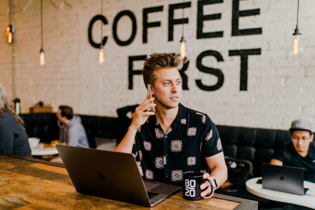 Ein Mann sitzt an einem Tisch in einem Café und hält ein Smartphone ans Ohr. Er blickt zur Seite und scheint auf etwas zu achten, während er ein Gespräch führt. Vor ihm auf dem Tisch stehen ein geöffnetes Laptop und eine Tasse mit dem Aufdruck "80 20". Im Hintergrund ist eine Wand mit der Aufschrift "COFFEE FIRST" und weitere Café-Besucher zu sehen.